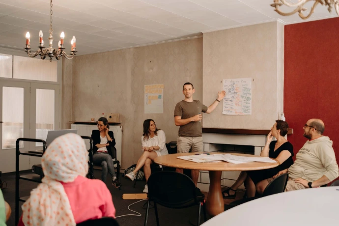Plusieurs personnes autour d'une table ronde sont assises et regardent un homme debout qui s'exprime